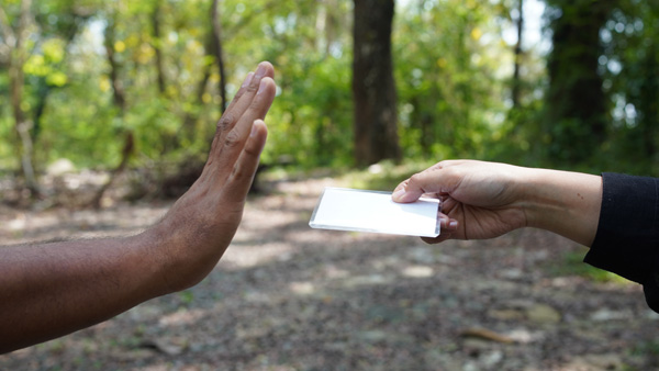 A hand is raised to refuse a blank card being offered by another hand in a wooded outdoor setting.