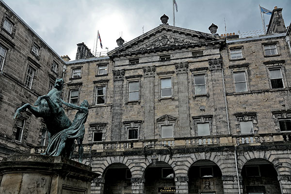 Historic stone civic building with arched entrances and ornate classical architecture (Edinburgh City Chambers), viewed from a courtyard with an equestrian statue in the foreground under an overcast sky.