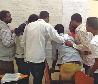 Group of seven people standing close together looking at 3 large sheets of hand written paper on a wall