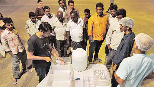 Group of people watching a man conducting an experiment on a table with boxes of equipment and a water bottle