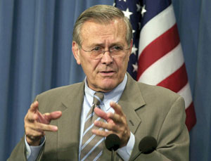 Photograph of Donald Rumsfeld speaking in front of microphones with the American flag behind him.