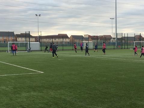 This image is of young people playing footbal informallyl on an artificial pitch. It is sunset and in the background there are houses.