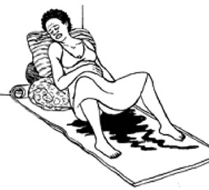 A woman is lying on a mat with her back and head supported by pillows. She is haemorrhaging and blood is covering the mat.