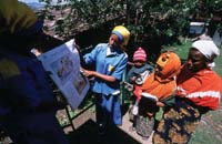 Three women are being shown hygiene education material by a sanitation guard. One of the women is holding a baby.