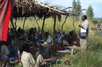 A community are gathered under a shelter listening to a health worker talk on health messages.