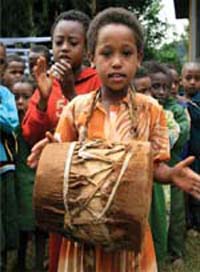 Children marching and clapping. A girl in the forefront playing a drum that hangs around her neck.