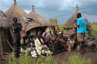 A health worker leading a discussion. A small group of people are sitting outside their homes together listening.