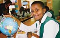 A student sits at their desk in a classroom. One hand rests on an open book and the other on a world globe on their desk.