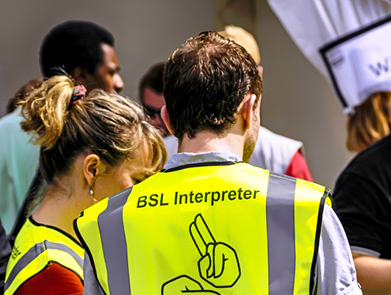 This image is a photo of two British Sign Language volunteers working at a public event. They are wearing high-vis yellow jackets with their backs to camera. On the back of the jackets, it says ‘BSL interpreter’ and shows a logo of a sign gesture. All around the people there are members of the public.