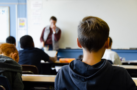 A photo of a child who looks to be in his early teens. The child is facing away from the camera and is sitting at a desk at the back of a classroom. Other students are around him. All the students are looking at a teacher standing at the front of the class.