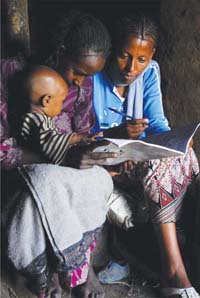 Two women a re looking through a workbook. A small child sits on one of the women’s lap.