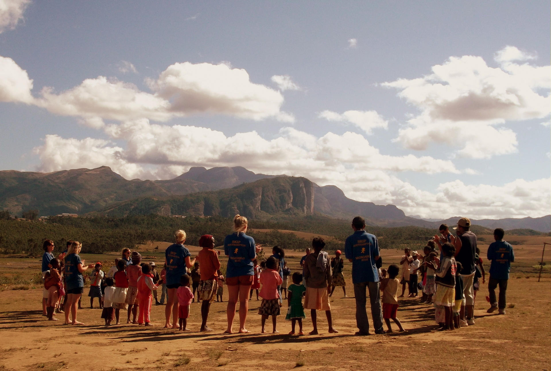 A group of SKIP volunteers stand in a circle with children, teaching them a game,They are stood in a desert, mountainous area