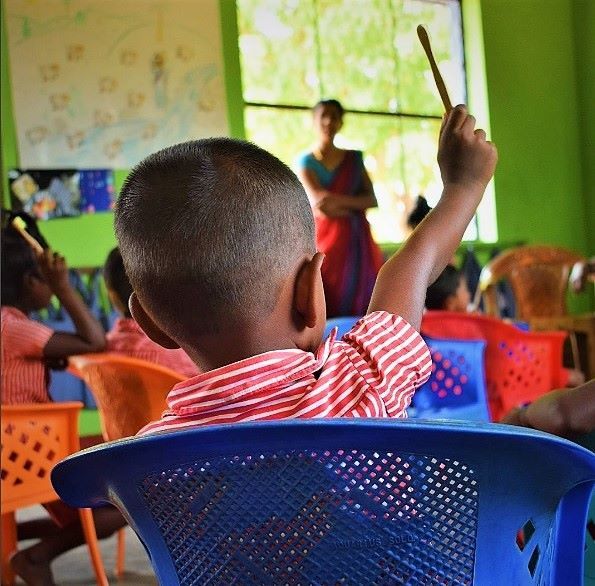 A child is sat in a green classroom facing a SKIP volunteer with their hand in the air holding a toothbrush