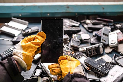 Photo of a person with gloves holding a broken smartphone surrounded by E-waste