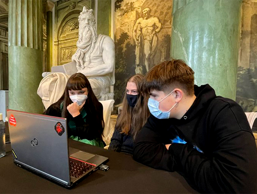 Troje uczniów siedzących przy stole patrzy w ekran laptopa