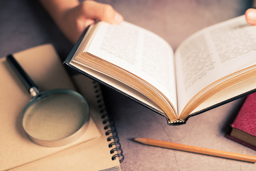 An image depicting a pair of hands holding an open book. Behind, on the desk, are a notepad, pencil and magnifying glass.
