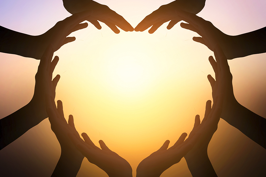 An image of several hands coming together to form a heart shape, which is backlit by a bright light.