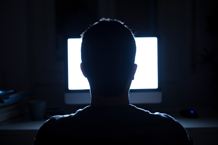 A silhouetted image of the back of a male head and shoulders. The person is in a dark room looking at a computer screen.