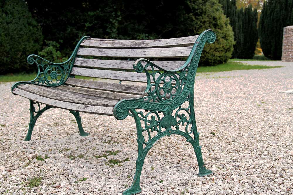 A rustic looking bench with weathered slats and green ironwork, sitting in an area of gravel surrounded by high trees.