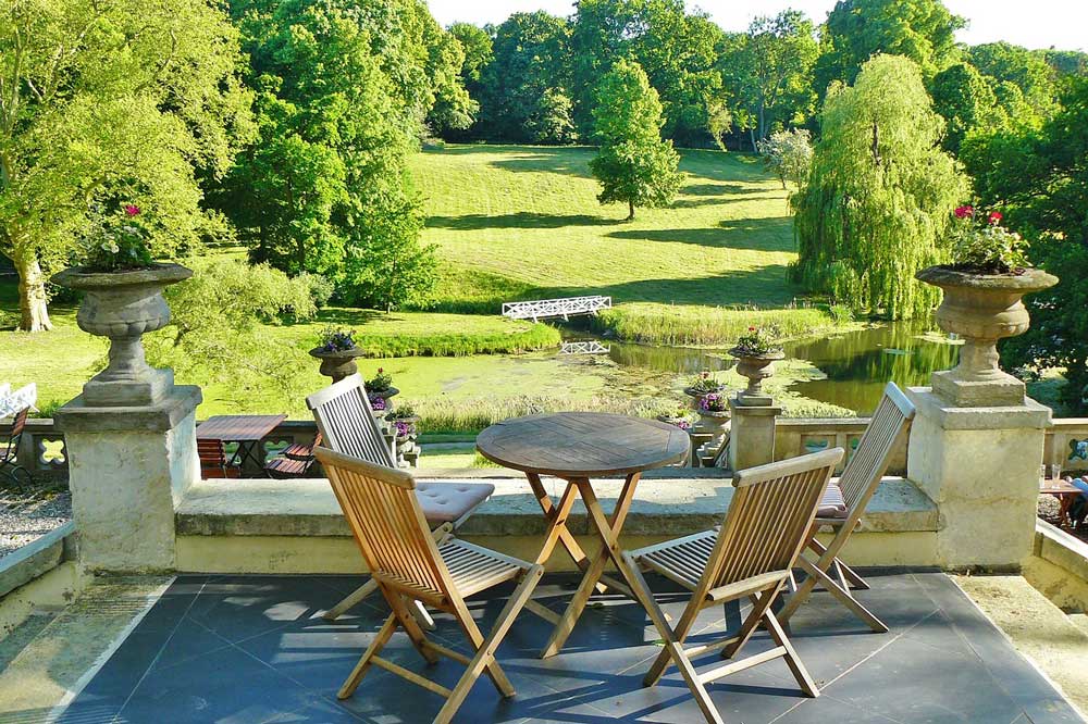 A wooden table and four chairs set out on a stone terrace overlooking green lawns surrounded by trees.