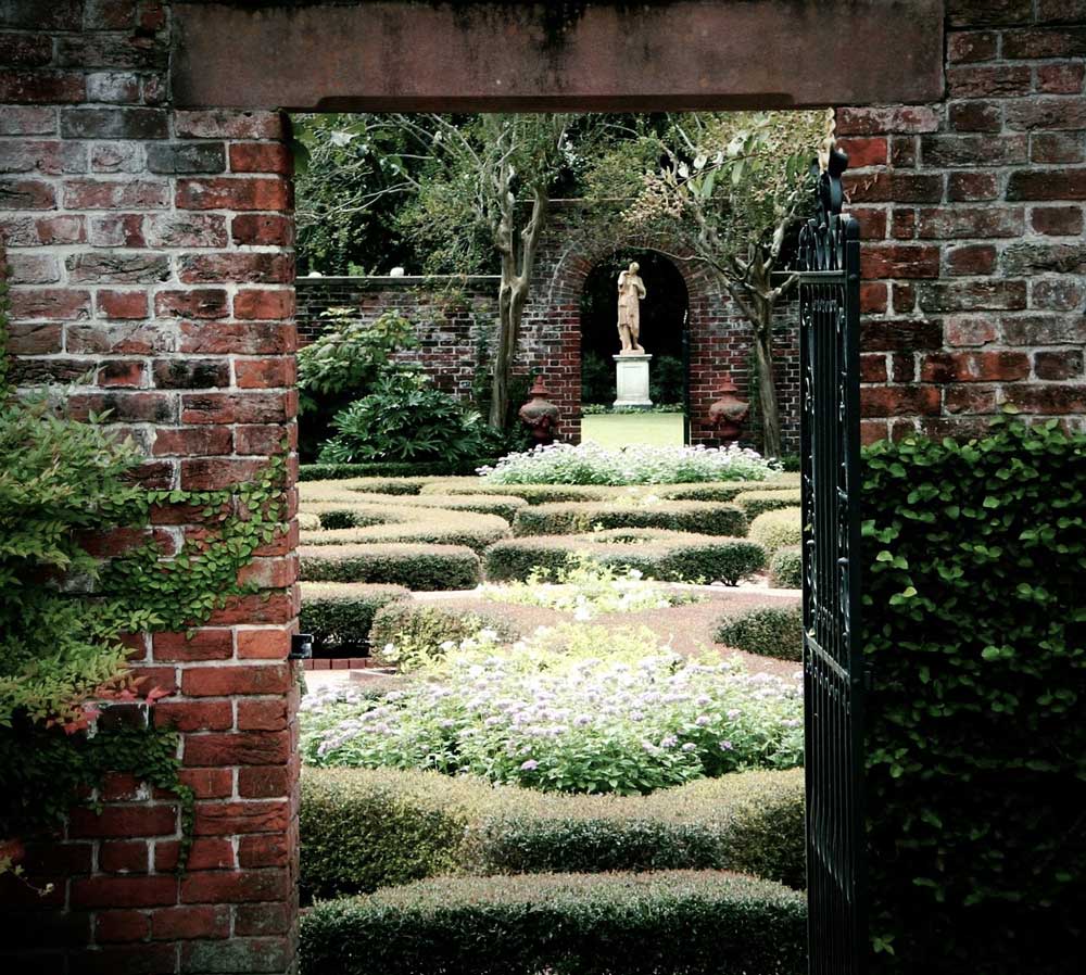A statue visible at the end of a formal garden through a brick archway.