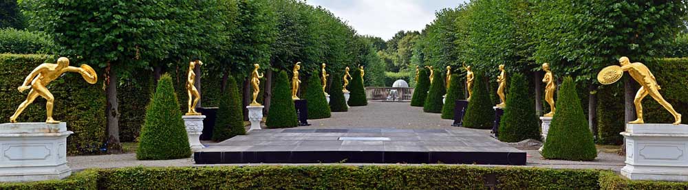 A path bordered by high hedges and topiary with evenly placed, gold coloured statues on plynths standing on either side.