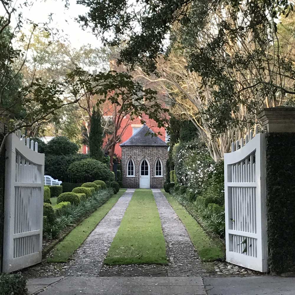 Open white gates onto a grass and gravel pathway leading to a small brick summerhouse with white arched windows and door.