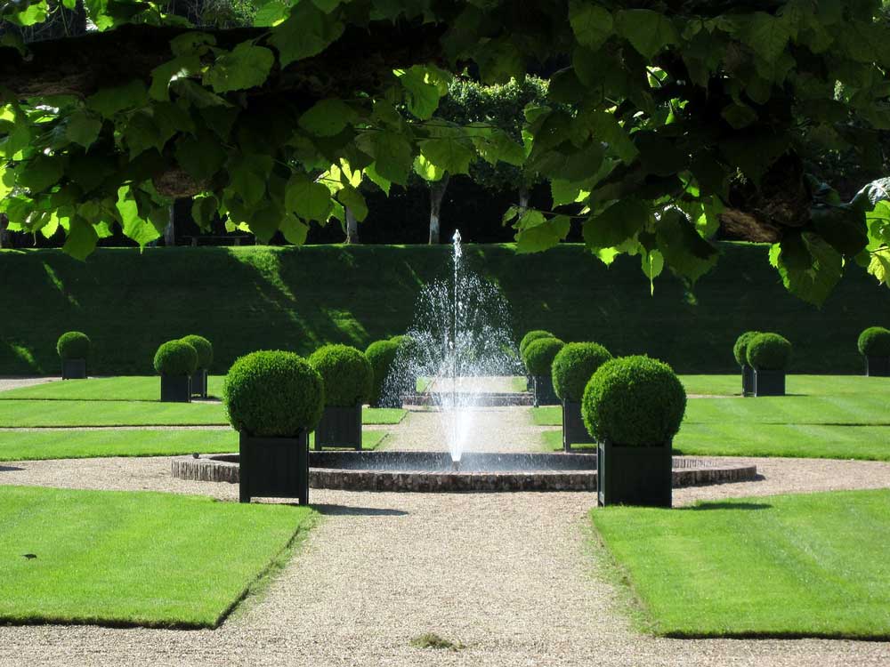 A formal garden with cut grass and round  topiary lining a path with a fountain in the centre.