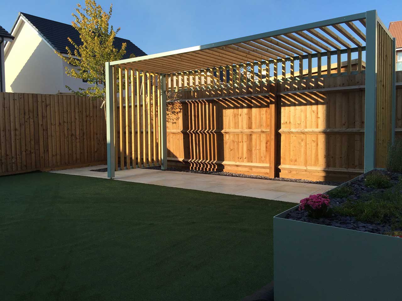 A slatted wood screen with two sides and a roof at the end of a garden catching sun.