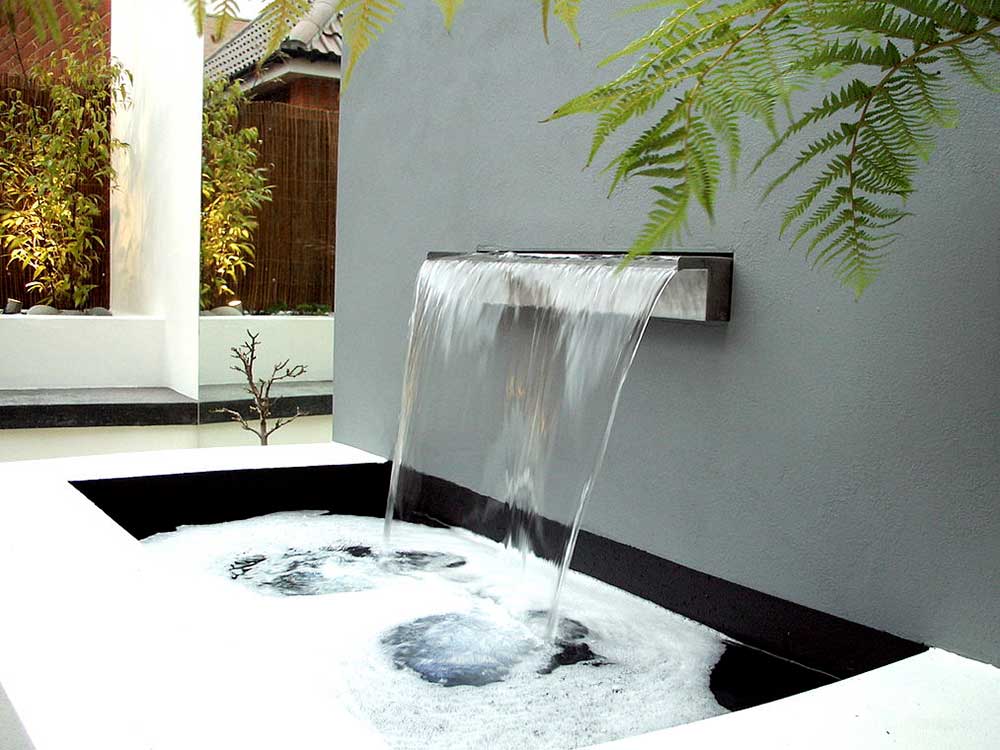 Modern garden with water flowing into a pool from an outlet on grey painted wall.