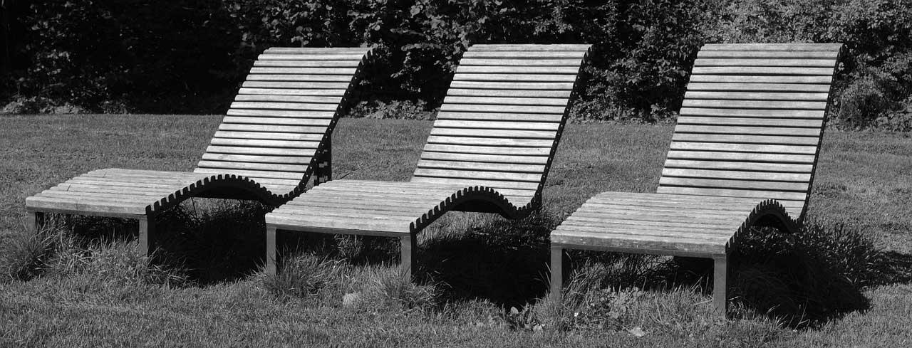 Black and white image of 3 wooden sunloungers side by side in a garden with hedging.