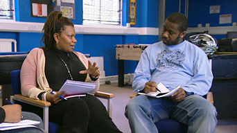 A photograph of a woman and a man sitting in an office environment, talking, each holding notes and a pen.