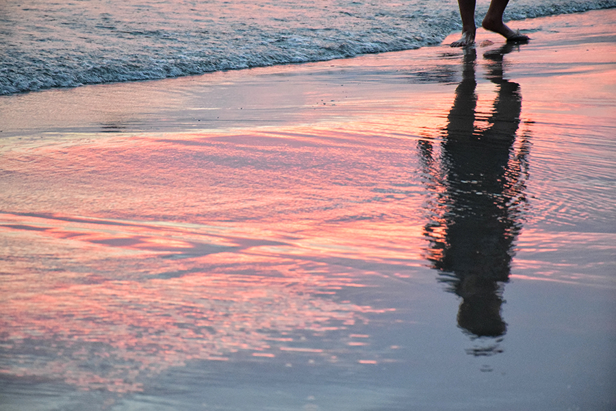 Une personne qui marche sur la plage, les pieds dans l’eau. L’image se concentre sur son ombre reflétée sur le sable humide.