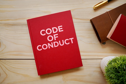 Un livre rouge intitulé « Code de conduite », une plante verte, deux autres livres et un stylo sur une table en bois.