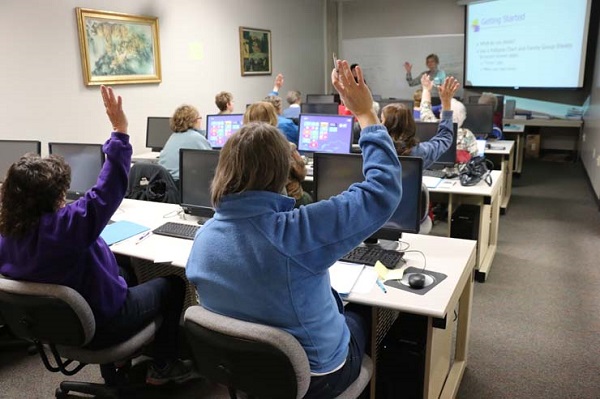 Students sitting behind computers with raised hands facing a facilitator at front of room.