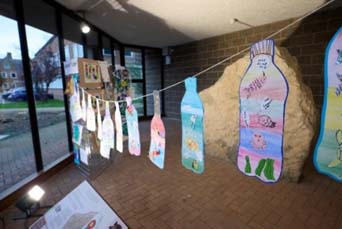 A length of string is hung across a room. On the piece of string are bottle shapes cut out from paper and decorated with different underwater scenes with speech bubbles next to different animals talking about their thoughts on human actions causing climate change
