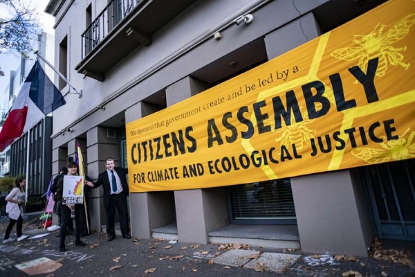 two people stand next to a large yellow banner on a building that has a French flag. The banner reads “We demand that government create and be led by a Citizens Assembly for climate and ecological justice”.