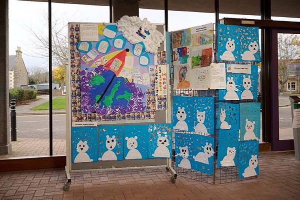 Display boards stand in an open space in front of large windows. A large hand-made poster featuring an image of the world covered with an umbrella and a cloud sits on the left-hand display board. There is a smaller poster on the right hand display board and both are surrounded by different collages of polar bears on blue backgrounds.