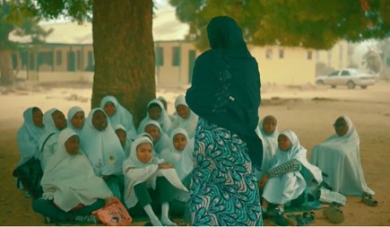 A group of female students sitting on the floor under a tree with the back of a teacher standing talking to them