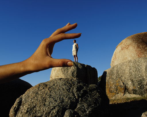 The image shows a person’s hand in the foreground, appearing very large, and a man standing on a rock in the background in the background, looking very small.