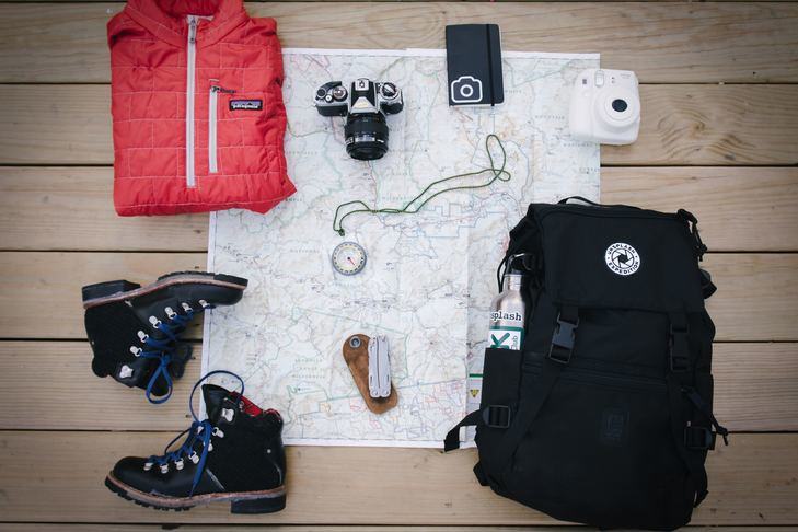 A photo of hiking items, including a map, boots, padded jacket, camera, compass, smartphone, rucksack and folding knife.