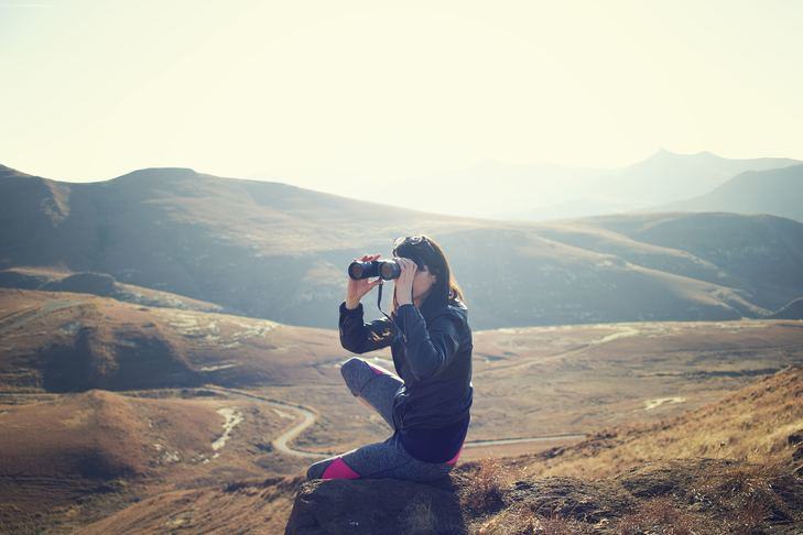 A photo of someone sitting on a small rocky outcrop on a brown hillside, looking into the distance using binoculars.