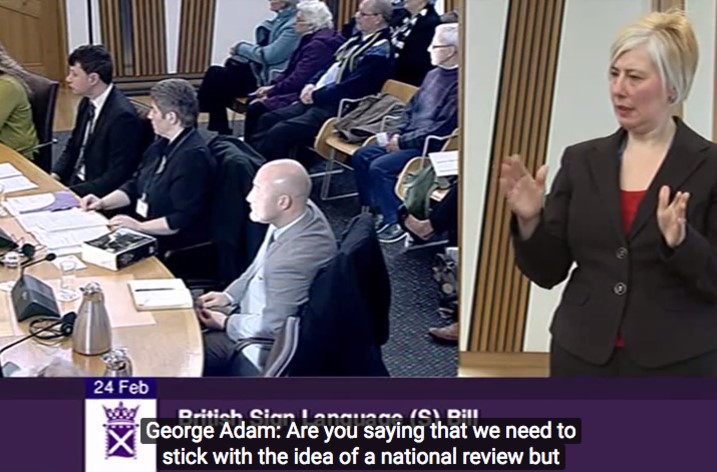 A screen capture of a debate in the Scottish Parliament, with separate panels showing a sign language interpreter & captions.