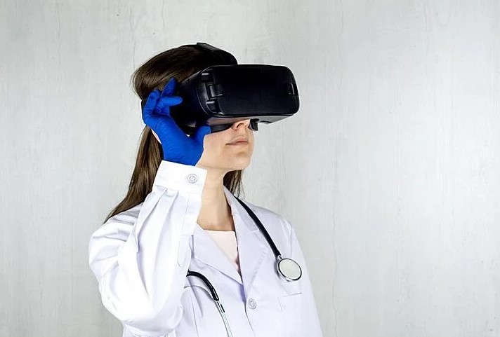 A photo of a person in a lab coat, latex gloves & with a stethoscope around their neck, wearing a VR headset over their eyes.
