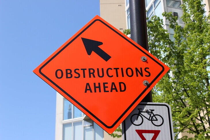 A bright orange diamond-shaped road sign with a black arrow pointing upwards and left and the words 'Obstructions ahead'. 