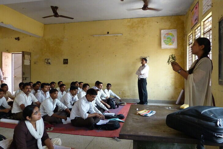 A classroom with adult students seated on the floor, genders separated. The teacher holds up a plant and she is speaking.