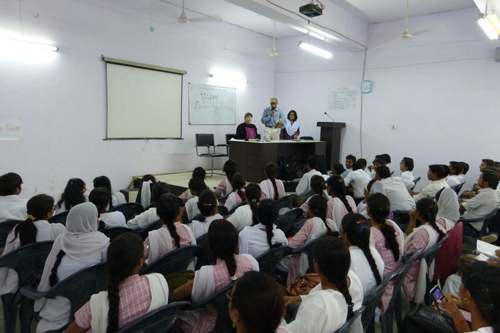 A large classroom with adult students seated in rows, gender-separated. 3 educators sit behind a desk, 1 speaking with a mic.