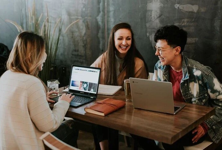 Three people sit around a table, two with open laptops and one a paper notebook. All are laughing. 