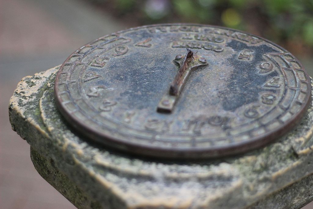 Close up of a round metal sundial with numbers and pointer, set on a stone pillar.