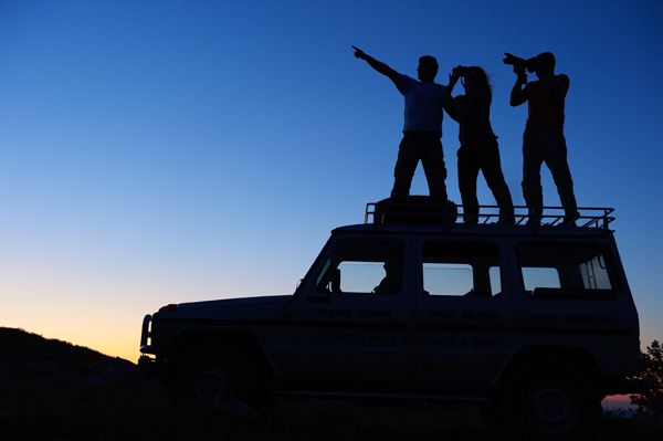 Three people standing on the roof of a vehicle, silhouetted against a twilight sky.  One person is pointing into the distance, another is looking through binoculars, and the third person appears to be adjusting a camera. The scene suggests exploration or adventure. The vehicle appears to be an off-road type, indicating they might be in a remote or rugged location.